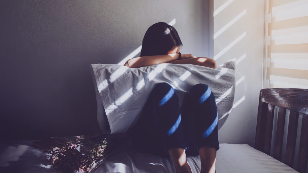 A woman is sitting hugging her knees in bed. Feeling sad.